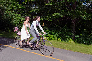 A bride and groom ride their tandem bike into the summer sunshine following their ceremony 