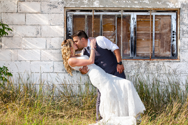 A groom dips and kisses his bride in front of an industrial building